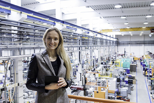 Horizontal color image portrait of blond business female worker standing on balcony on top of large factory, holding digital tablet and examining the production online. Focus on attractive businesswoman sincerely smiling at camera, futuristic machines in background.