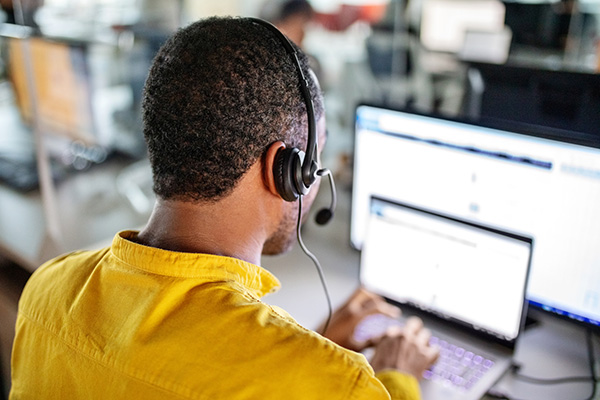 Rear view of a mature man with a headset sitting at the desk working on a laptop. Male customer service representative working in the office.